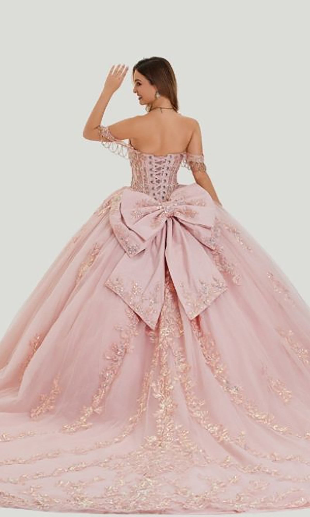 A woman faces away and waves while wearing the Quinceanera Dress 26409 by Stella Couture, a pastel pink ball gown with embroidered details and a large bow on the back.